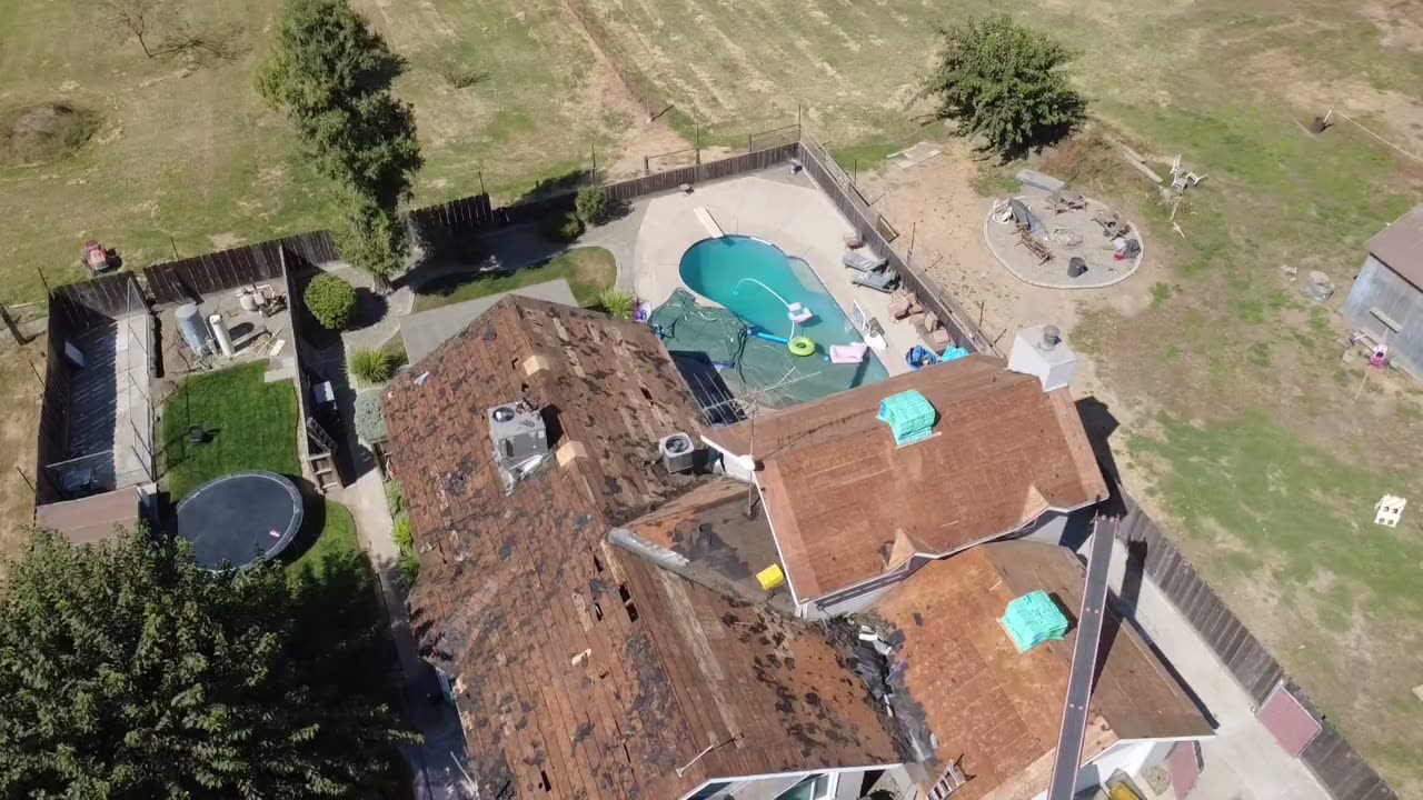 Drone view of a house mid tear-off: old shingles stripped, pool and dumpster below